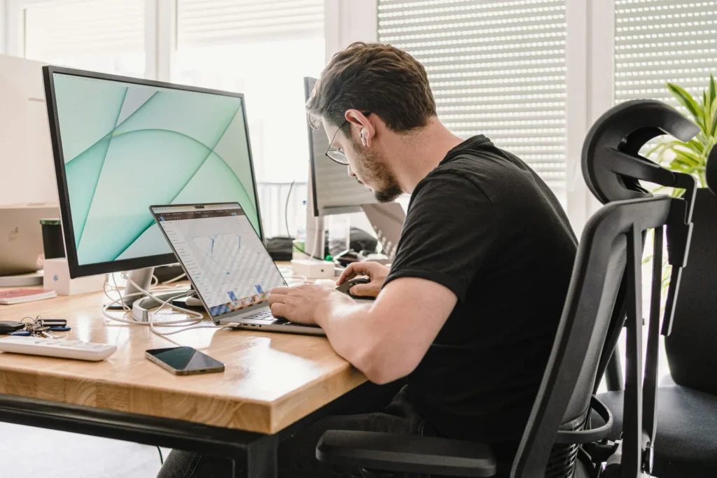 Man at a desk looking at a laptop next to a desktop monitor