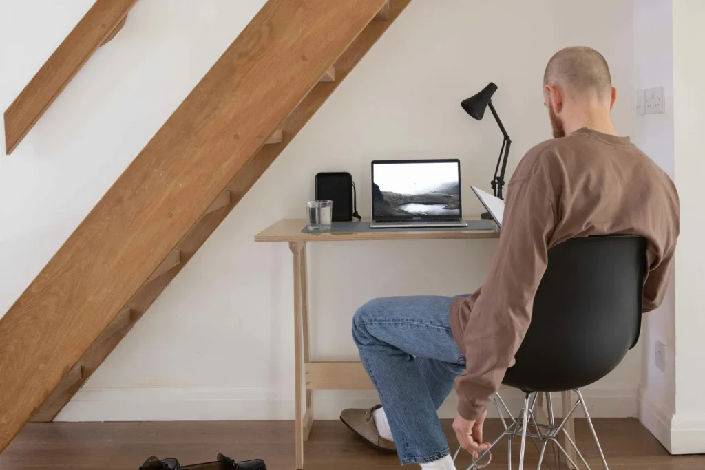 Man seated at a small desk with a laptop and lamp under a staircase