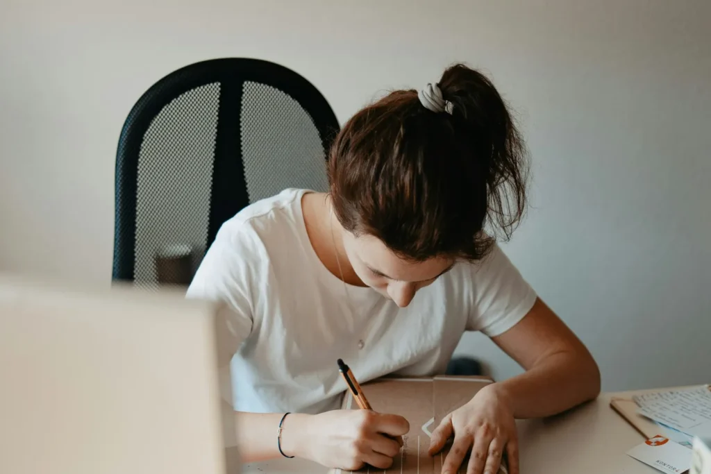 Person writing notes in a notebook at a desk next to a laptop