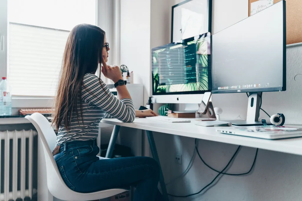 Woman at a desk with two monitors, resting her chin on her hand