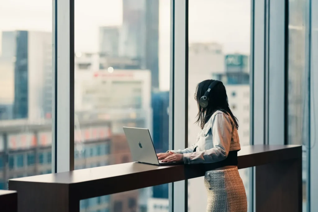 Woman with headphones using a laptop at a counter by a city window